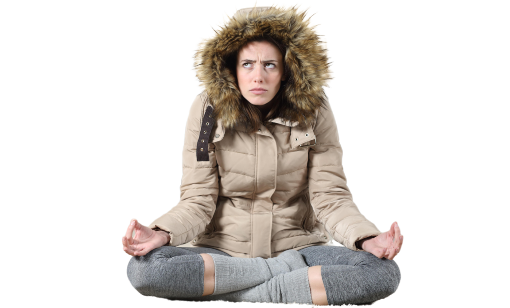 Woman wearing a beige puffer jacket with fur-lined hood and gray leggings sits cross-legged against a white background, appearing cold and uncomfortable.
