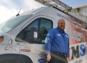 Smiling HVAC technician in blue uniform standing beside white service van with Carrier branding and contact information.